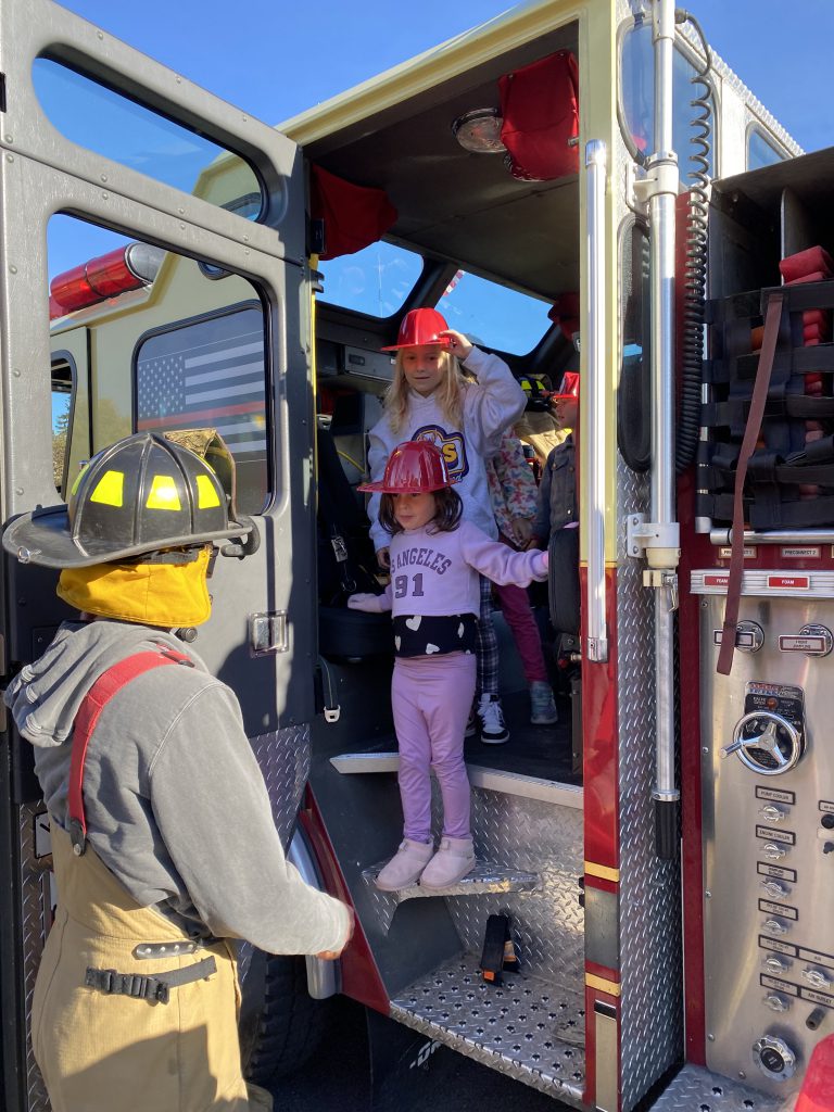 Two elementary students wearing red fire helmets stand inside a fire truck while a firefighter assists them down the stairs.