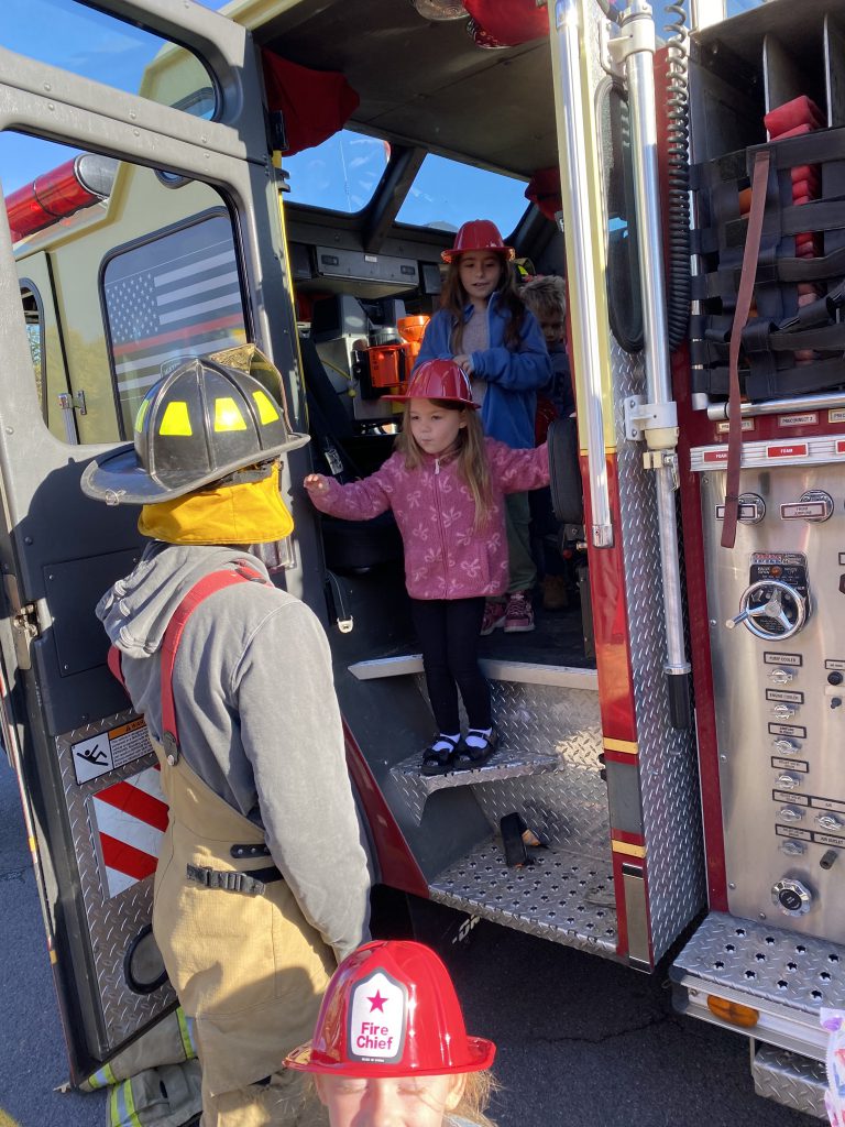 Two elementary students wearing red fire helmets stand inside a fire truck while a firefighter assists them down the stairs.