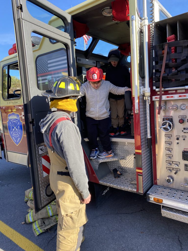 Two elementary students wearing red fire helmets stand inside a fire truck while a firefighter assists them down the stairs.