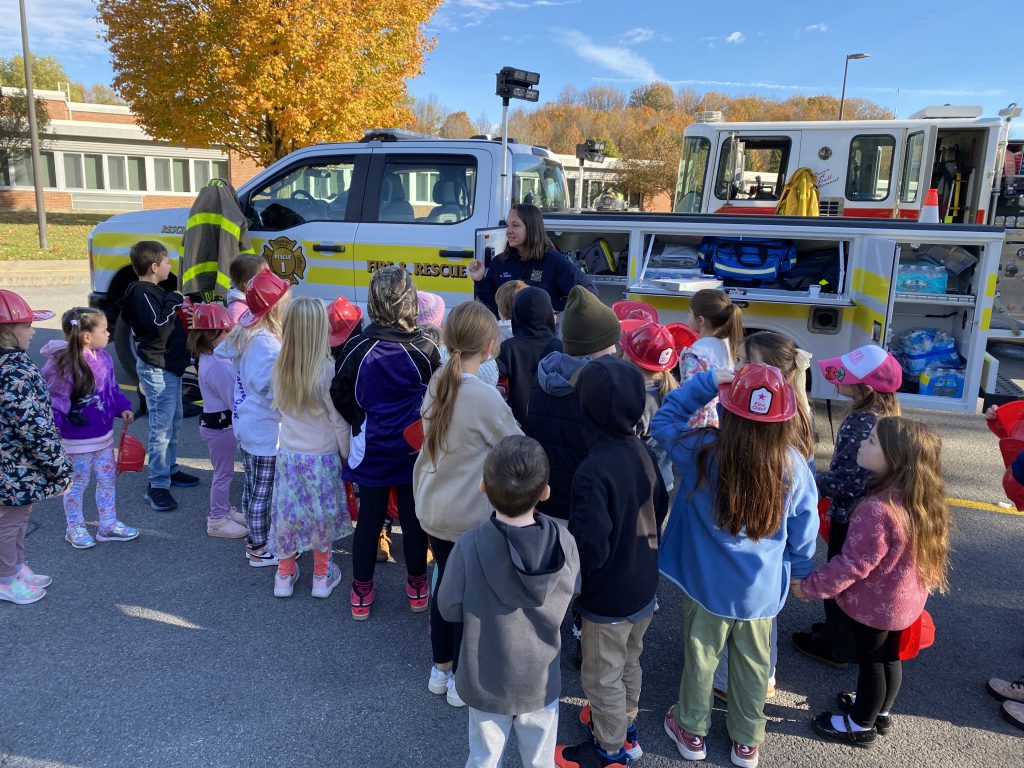 A group of students gathers around a fire and rescue truck as a volunteer firefighter explains its features and equipment.