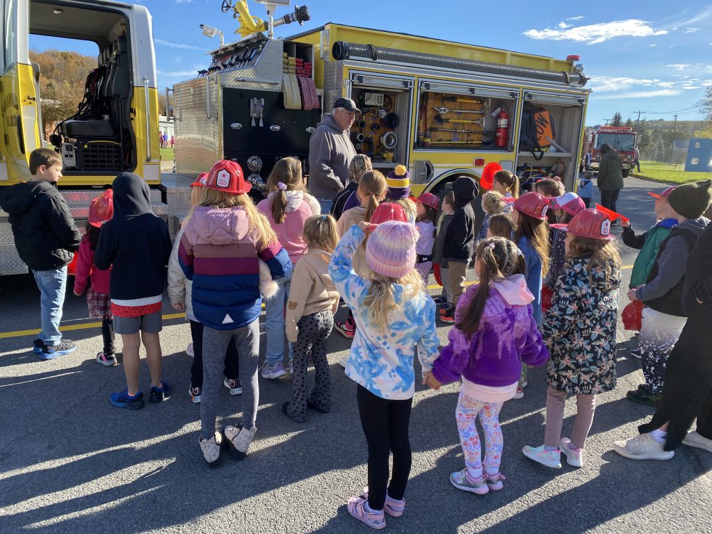A group of students gathers around a fire and rescue truck as a volunteer firefighter explains its features and equipment.