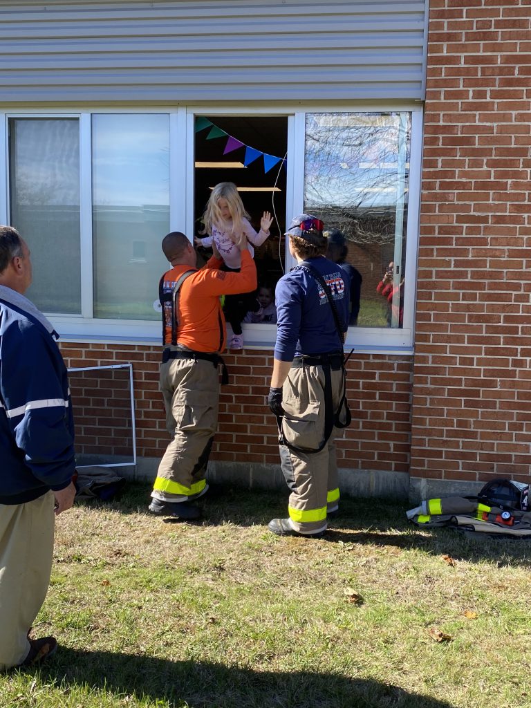Two firefighters assist a student as they climb out of a window during a practice fire drill.