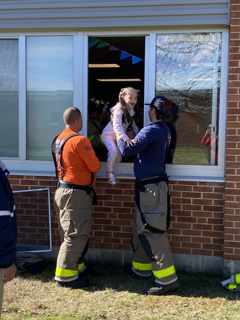 Two firefighters assist a student as they climb out of a window during a practice fire drill.
