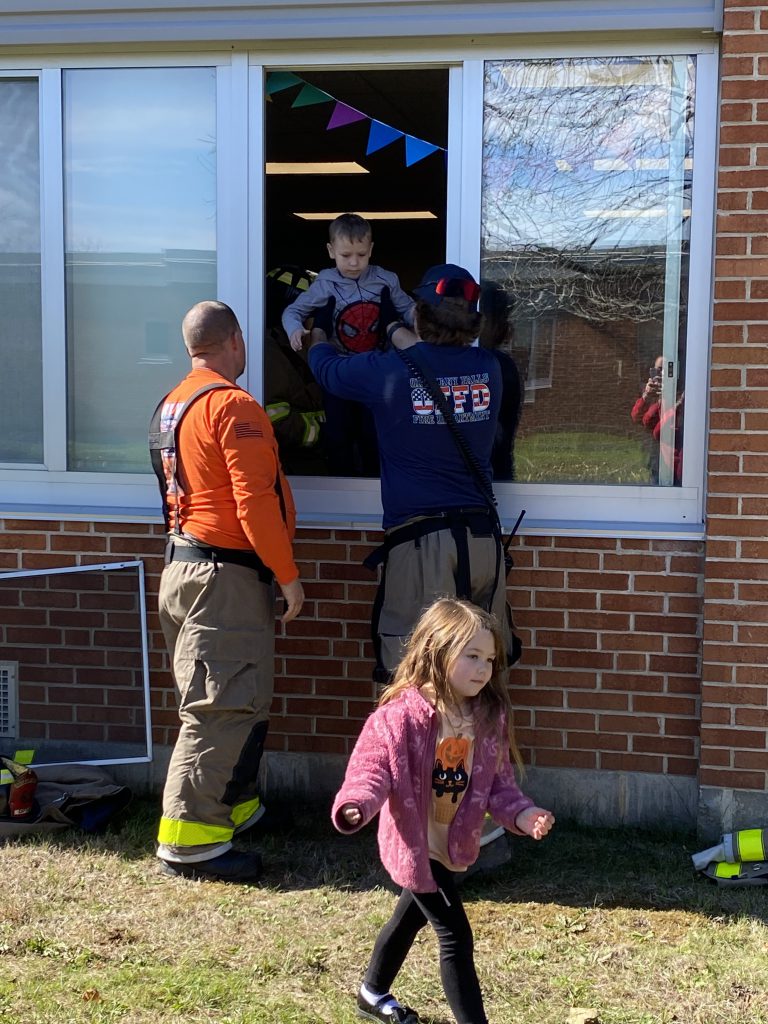 Two firefighters assist a student as they climb out of a window during a practice fire drill.