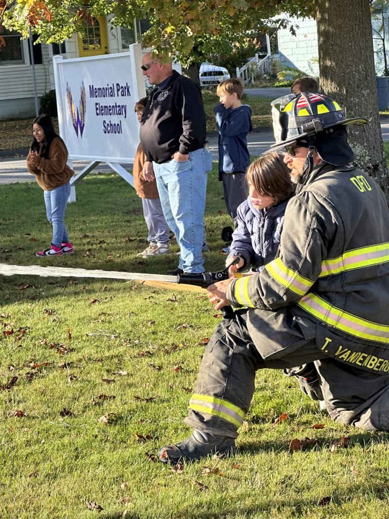 A firefighter assists a student in holding and aiming a fire hose during a safety demonstration.