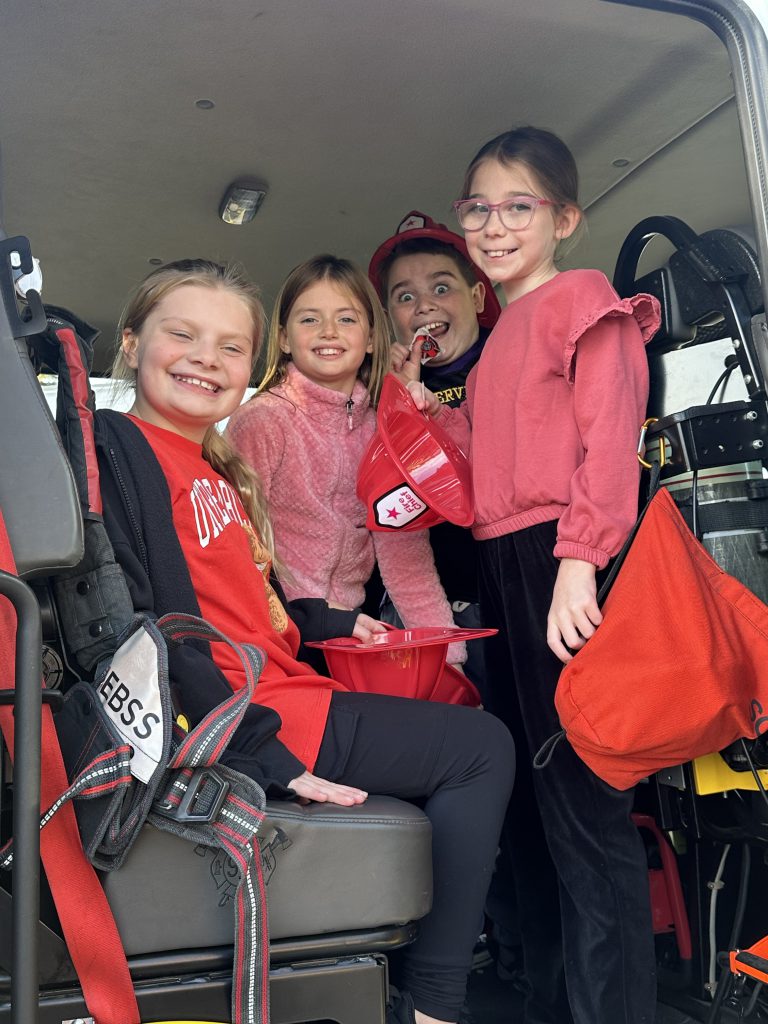 Four students smile and pose for a photo while sitting inside a firetruck during a school visit.