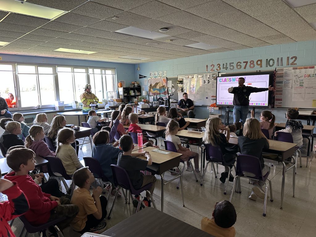 A firefighter gives a safety presentation to a classroom full of elementary students, sharing fire prevention tips and demonstrating equipment as part of Fire Safety Week activities.