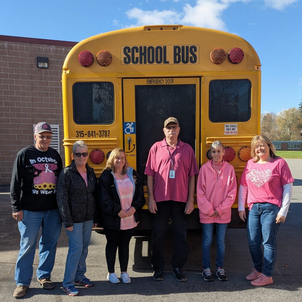 six employees of the bus garage are pictured in front of a school wearing pink