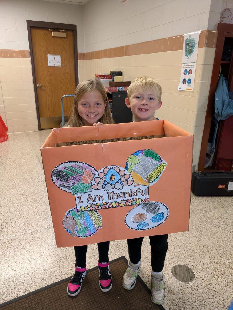 An elementary school girl and boy hold an orange cardboard box decorated with a hand-drawn turkey, food items, and the words “I am thankful.”