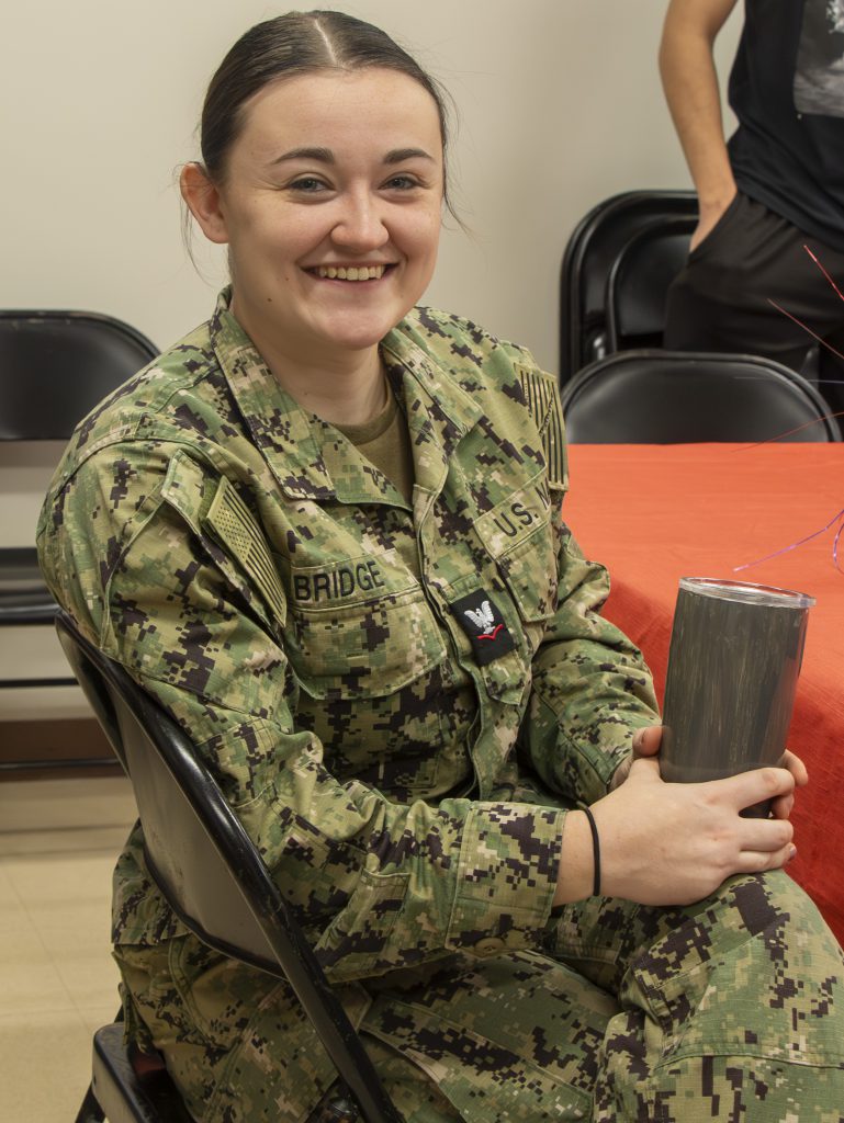 A woman soldier in an army uniform sits on a chair, holding a travel mug in both hands, while smiling.