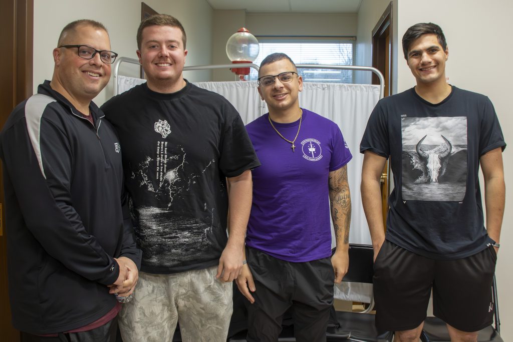 Four men standing side by side, smiling at the camera; three wear black t-shirts and one wears a purple t-shirt.