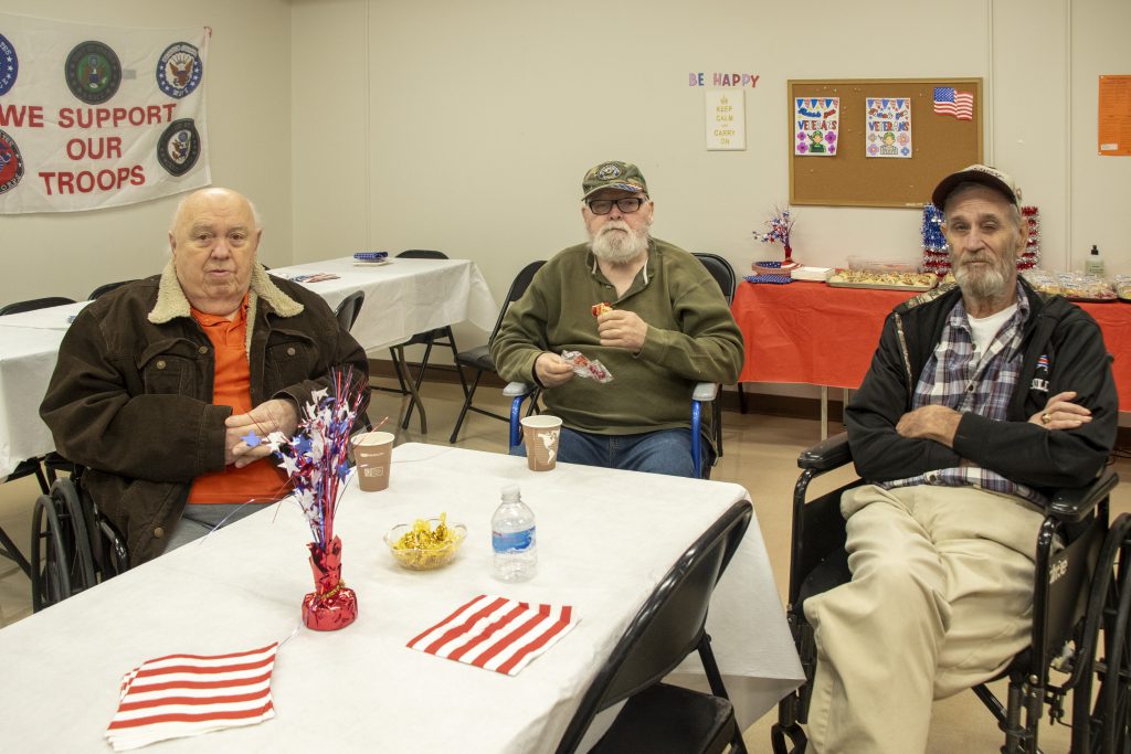 Three retired veterans sitting together at a table covered with a white tablecloth, decorated with red, white, blue accents; each has a coffee cup in front of them.