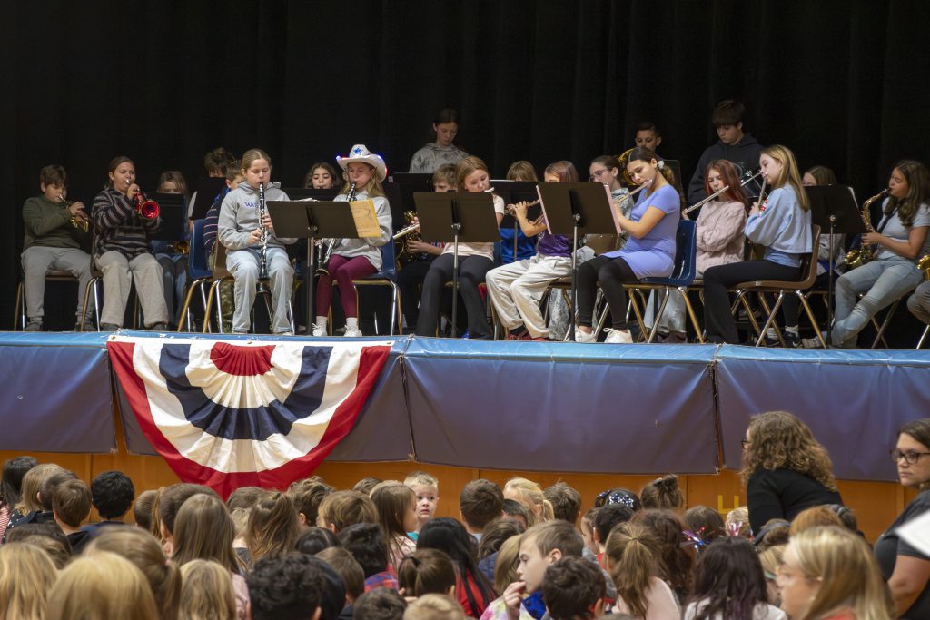 Elementary school students performing in a band on stage, playing instruments on a stage decorated in red, white and blue. 