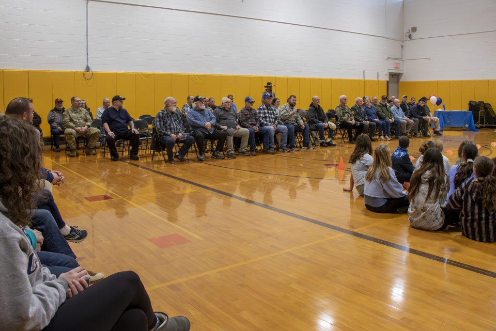 Group of veterans seated together in a school gymnasium, gathered for a veterans day celebration. 