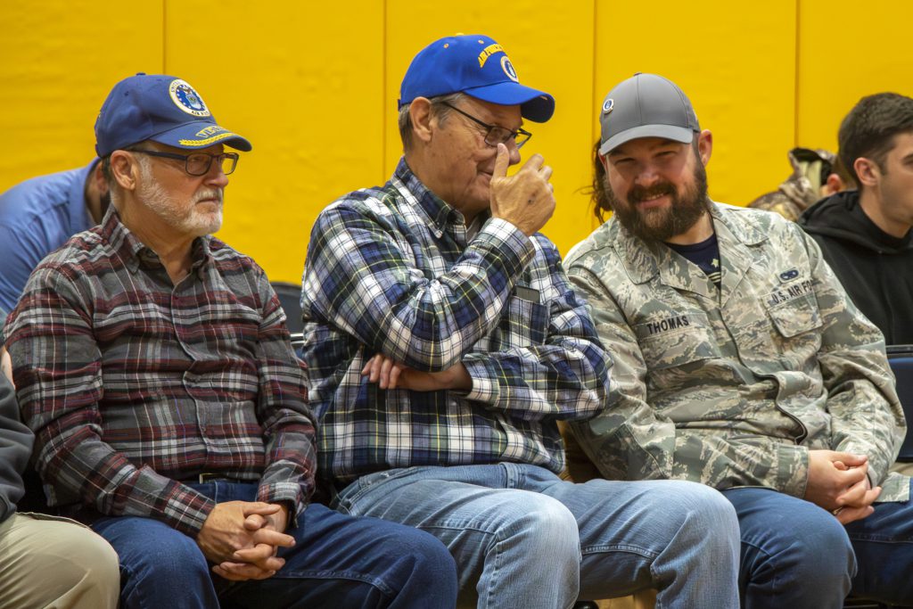 Three retired veterans sitting together engaged in conversation; the man in the middle gestures with one hand raised near his face while speaking and the man on the right is smiling.