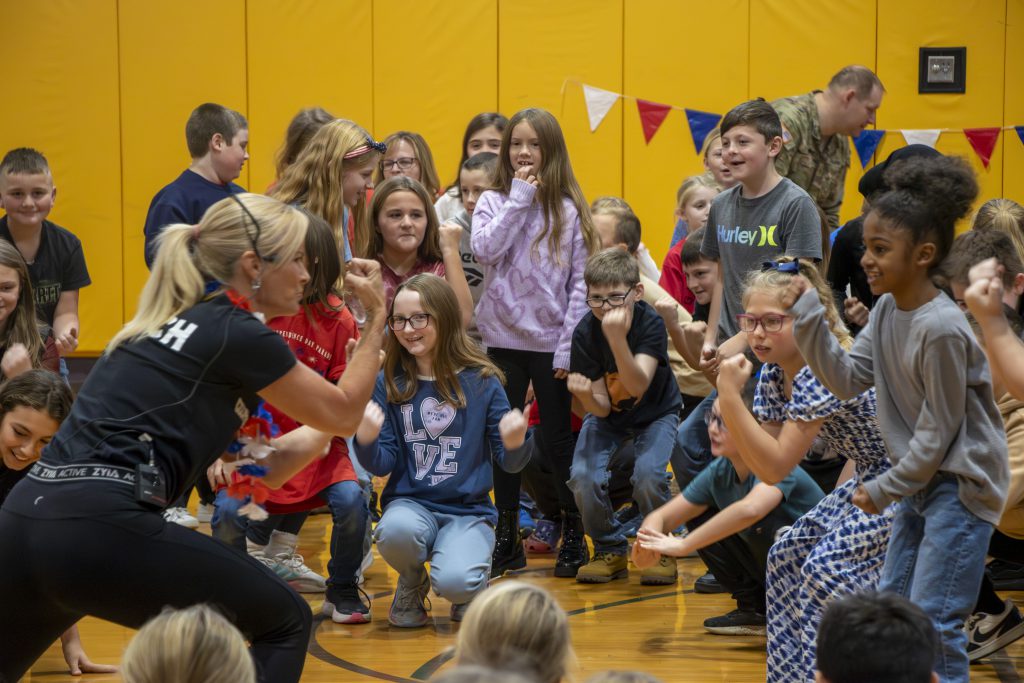 coach Lisa Briggs, wearing a black t-shirt labeled 'Coach,' leads elementary students in a boot camp challenge during a school event honoring veterans with physical activities and tributes. 