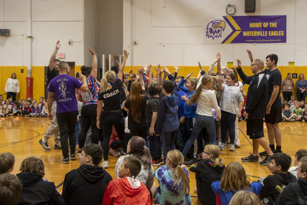 Coach Lisa Briggs, wearing a black t-shirt labeled 'Coach,' leads elementary students in a boot camp challenge during a school event honoring veterans with physical activities and tributes.