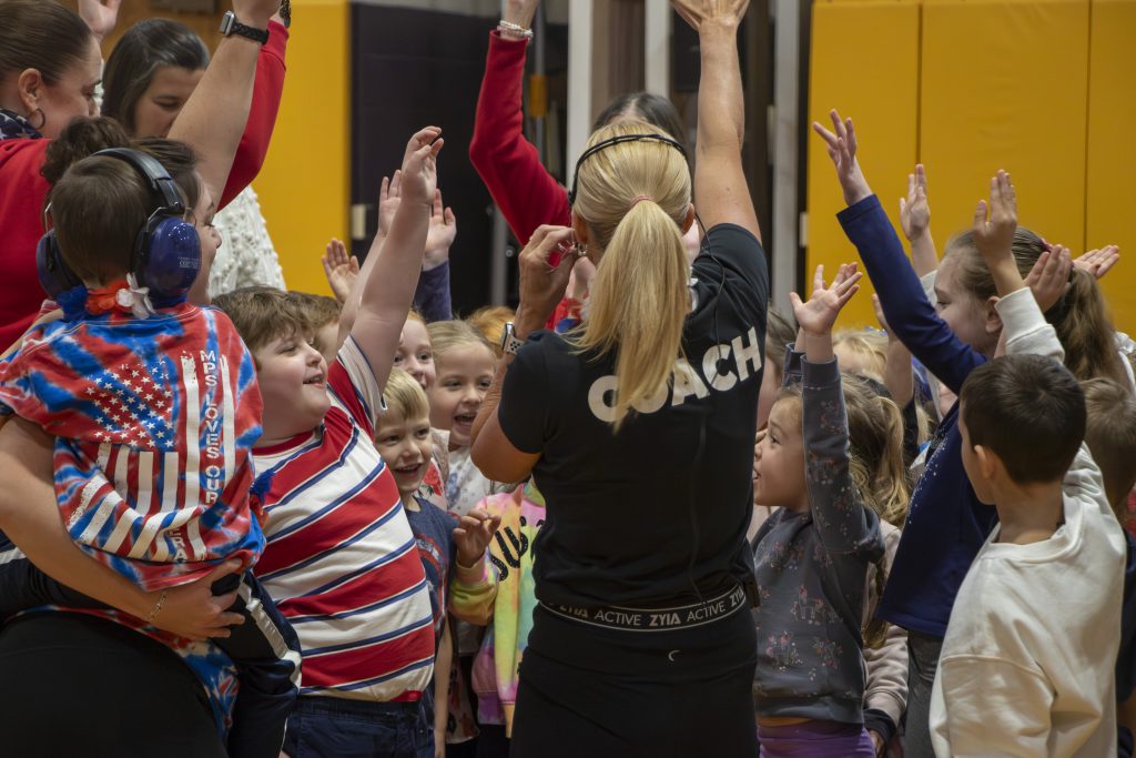Coach Lisa Briggs, wearing a black t-shirt labeled 'Coach,' leads elementary students in a boot camp challenge during a school event honoring veterans with physical activities and tributes.
