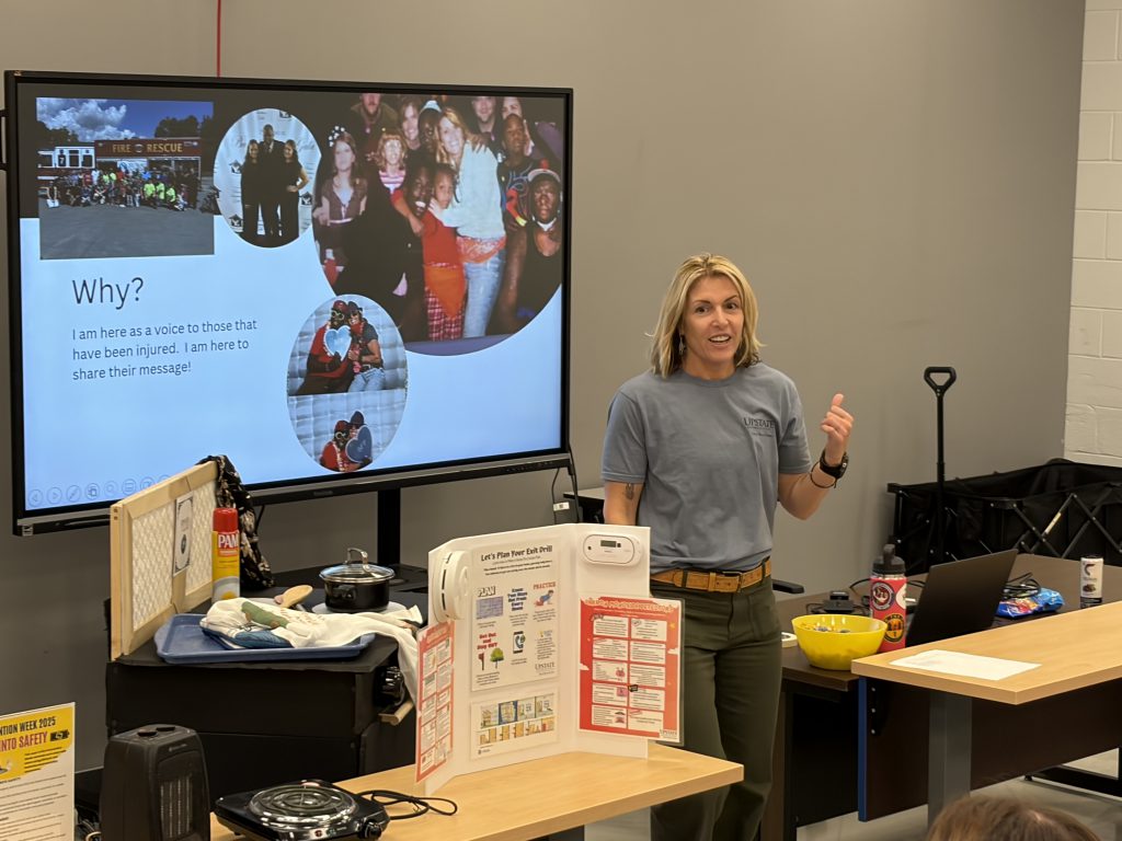 The image shows a woman standing in front of a large projection screen displaying various images, surrounded by various items on a table in the foreground.