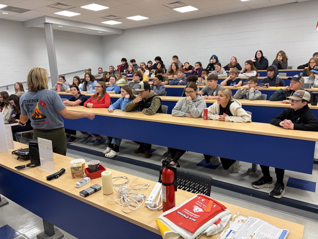 A crowded classroom with students seated at desks, some holding books or other materials, and a person standing at the front of the room.