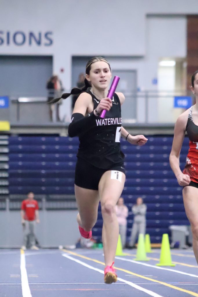 A female runner in athletic gear strides forward on an indoor track, carrying a purple baton in her right hand, her posture upright and focused, with lane markings visible beneath her feet and the curved track stretching ahead.