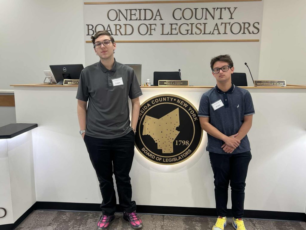 Two high school students standing in front of a sign that reads "Oneida County Board of Legislators" alongside a gold sign of Oneida County, New York, including an outline of the county map that has a date of 1798 and reads Board of Legislators.