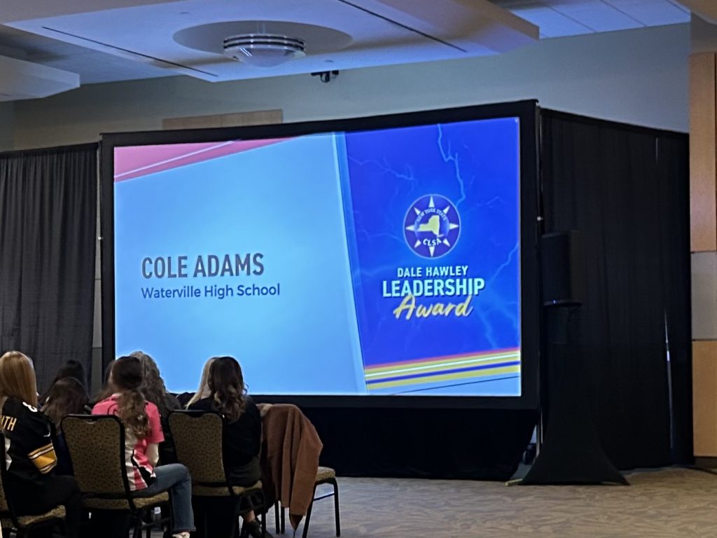 The image shows a large projection screen displaying the "Cole Adams Leesville High School Leadership Award" against a backdrop of a dimly lit room with people seated in chairs.
