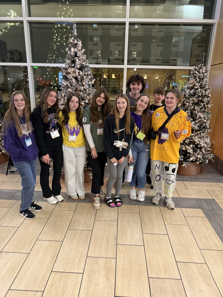 A group of young people, likely students, posing together in front of a decorated Christmas tree in what appears to be a lobby or entrance area of a building.