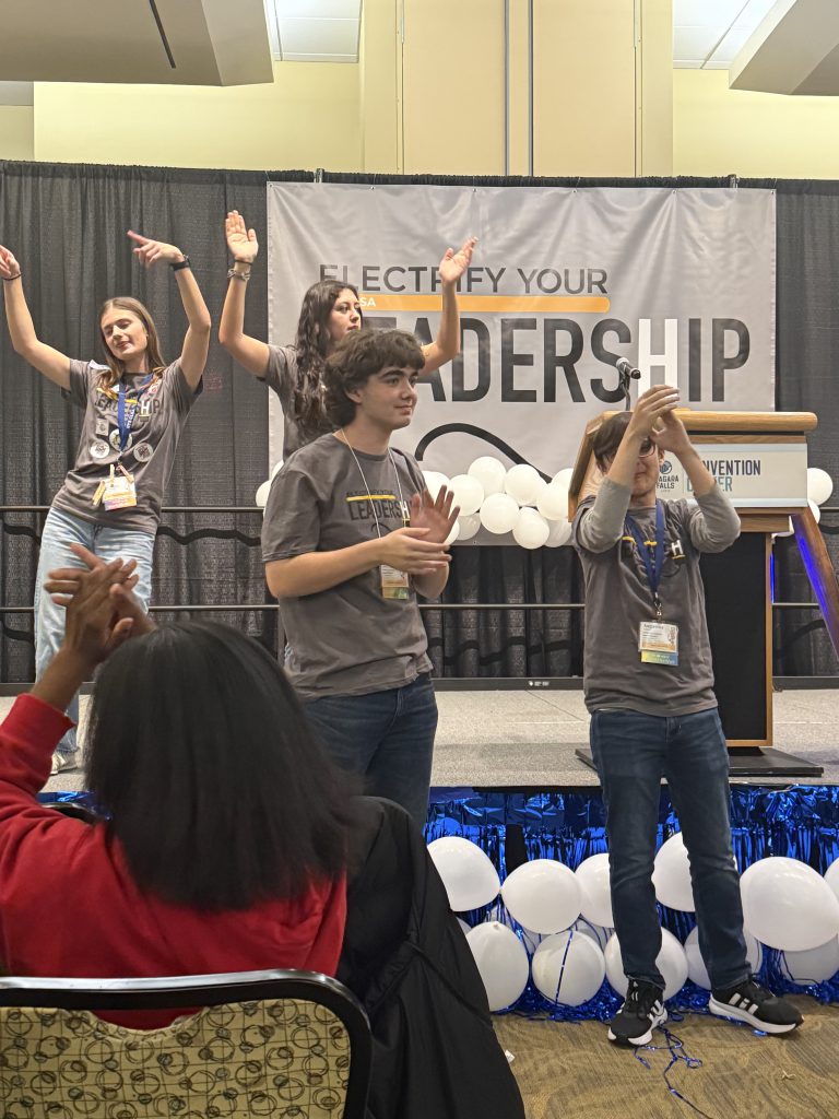 The image shows a group of people on a stage, with a banner in the background that says "Electrify Your Leadership". The people appear to be engaged in some kind of presentation or performance, with their arms raised and gesturing.