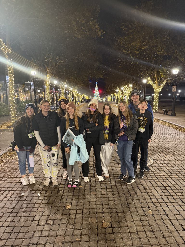A group of young people, likely students, posing together in front of a decorated Christmas tree in what appears to be a lobby or entrance area of a building.