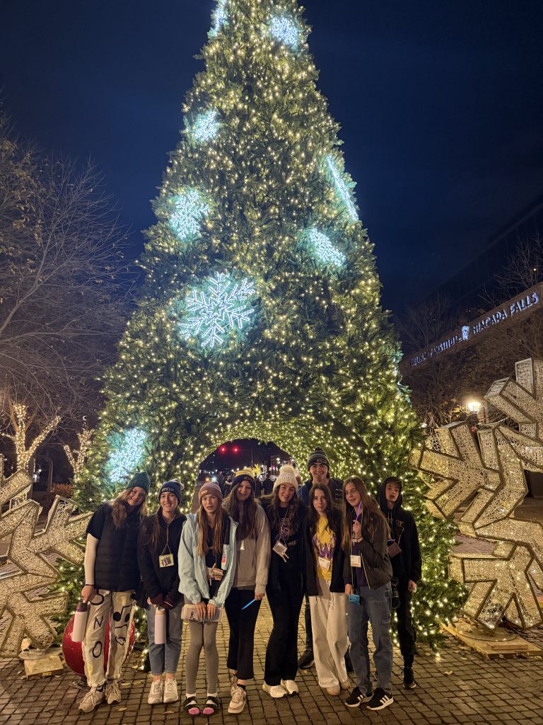 A large, brightly lit Christmas tree stands in the foreground, surrounded by a group of people posing for a photograph in the dimly lit outdoor setting.