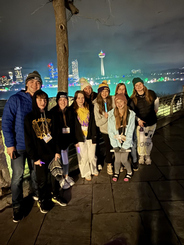 A group of people, likely friends or family, posing together in front of a nighttime cityscape with a tall tower and colorful lights in the background.