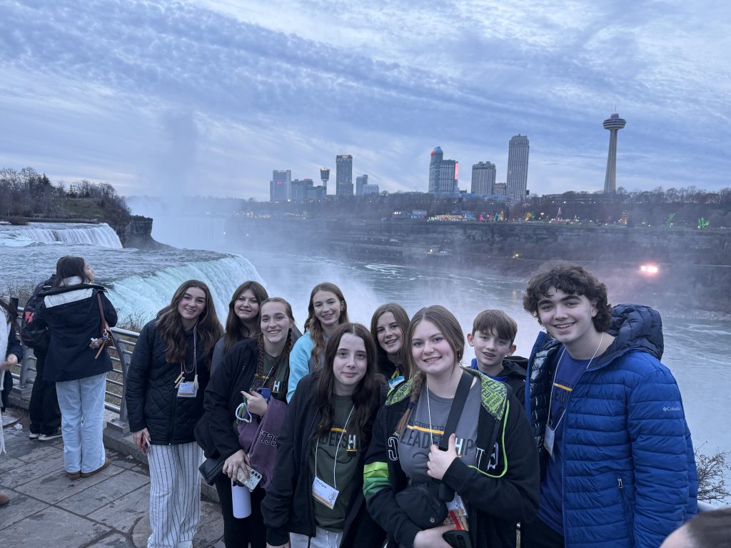 A group of people, likely a school or youth group, standing in front of the majestic Niagara Falls, with the city skyline of Niagara Falls, Ontario visible in the background.