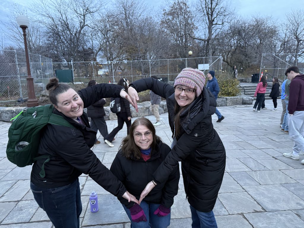 The image shows three young women standing together outdoors on a paved surface, with trees and buildings visible in the background.