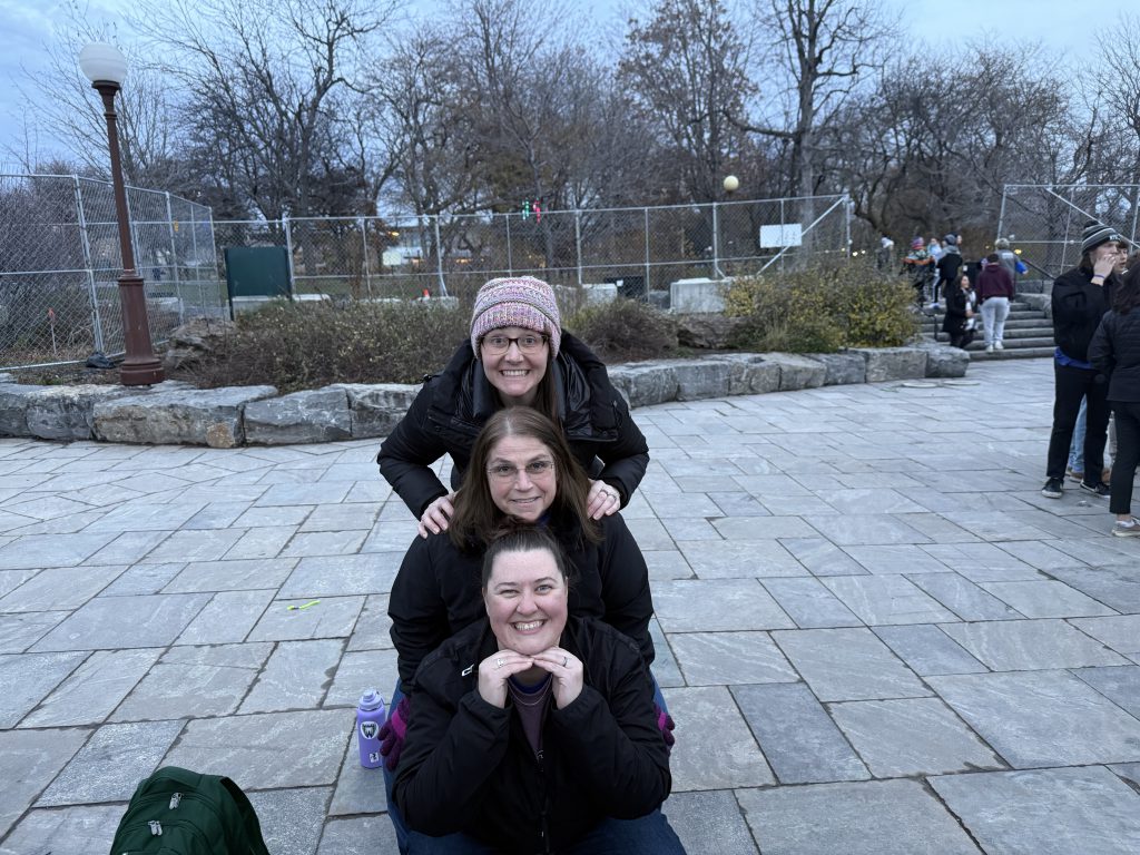The image shows three people, standing together outdoors on a paved walkway surrounded by trees and other vegetation.