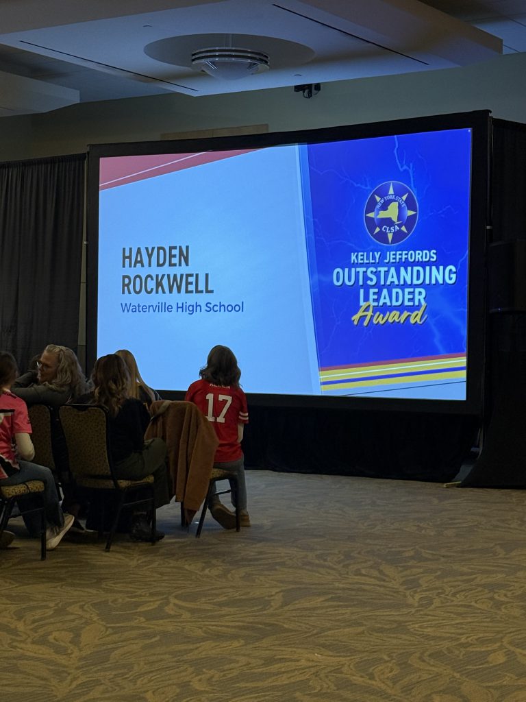 The image shows a large projection screen displaying the name "Hayden Rockwell" and the text "Waterville High School" along with the "Kelly/Jeffords Outstanding Leader Award" logo. In the foreground, there are several people seated in chairs, suggesting this is likely an award ceremony or presentation.