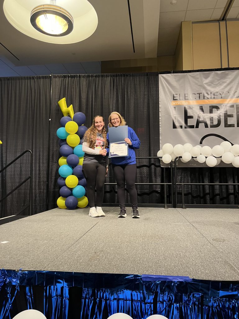 The image shows two women sitting on a stage, surrounded by colorful balloons and a backdrop with the text "Brittney Leader". The stage appears to be set up for some kind of event or presentation.