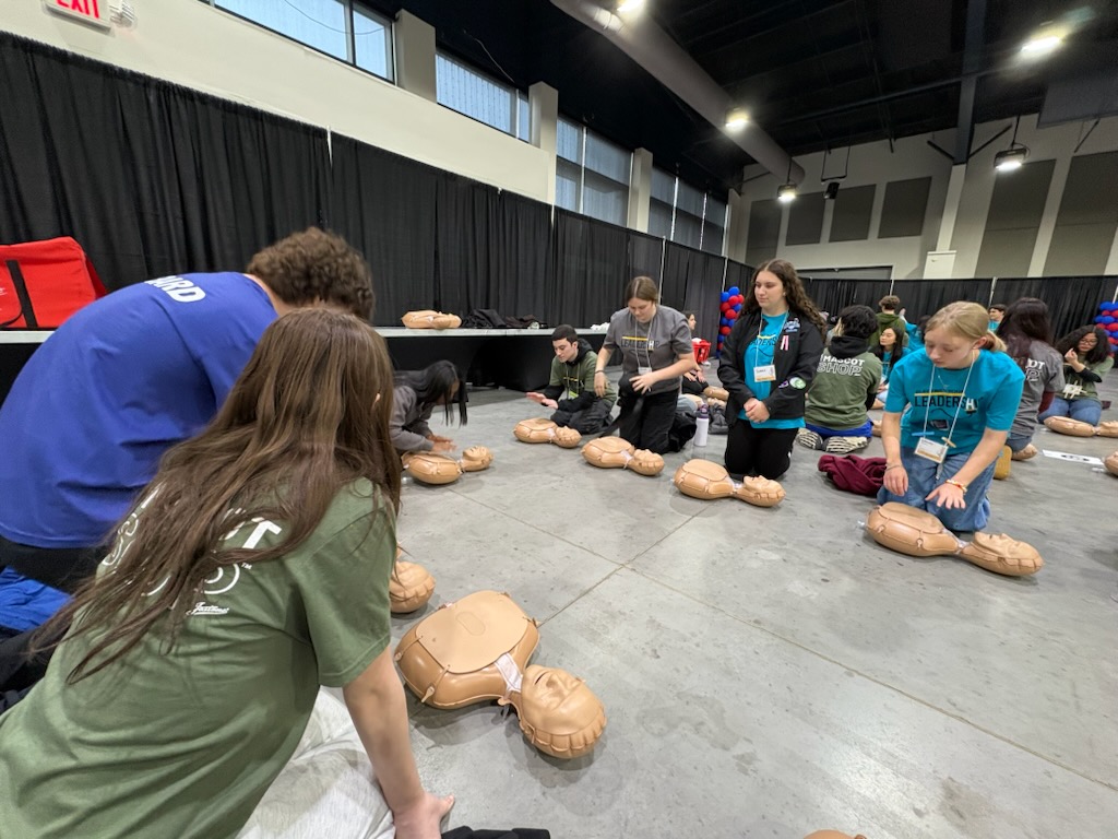 A group of people, likely attending a conference or event, are gathered around several CPR training mannequins on the floor, practicing lifesaving techniques in a large indoor space with curtains and equipment visible in the background.