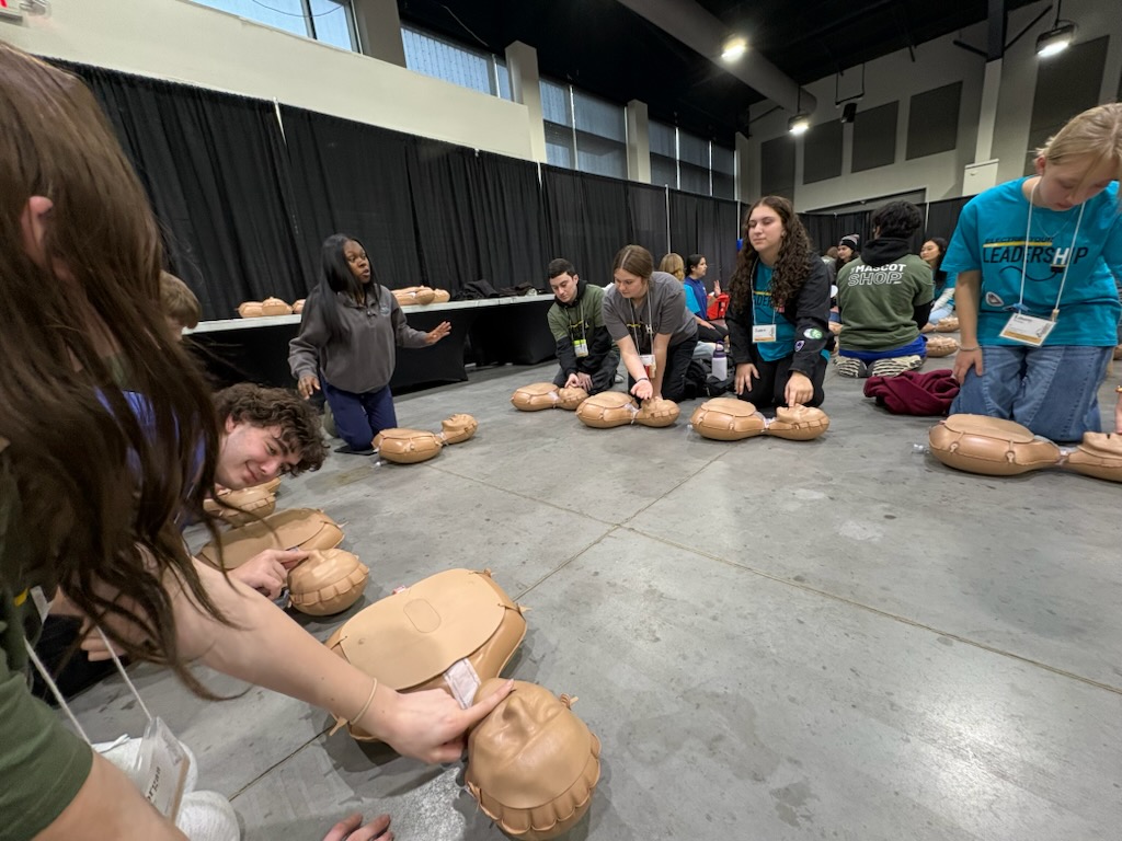 A group of people, likely attending a conference or event, are gathered around several CPR training mannequins on the floor, practicing lifesaving techniques in a large indoor space with curtains and equipment visible in the background.
